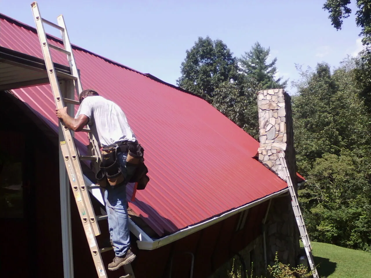 Roofer on a ladder beside a red metal roof and stone chimney — Soffit & Fascia Repair by Roofing Brothers in Thatcher
